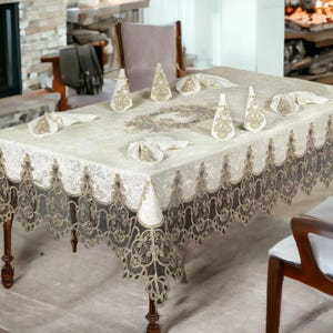 May include: A rectangular cream-colored tablecloth with a decorative lace border and matching napkins and decorative tree-shaped items. The table is set for a formal occasion, with a fireplace in the background.