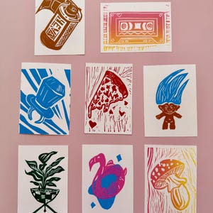 May include: A collection of linocut prints on white paper. The prints feature various designs, including a film canister, a cassette tape, a ring, a pizza slice, a troll doll, a potted plant, a flamingo float, and a mushroom.