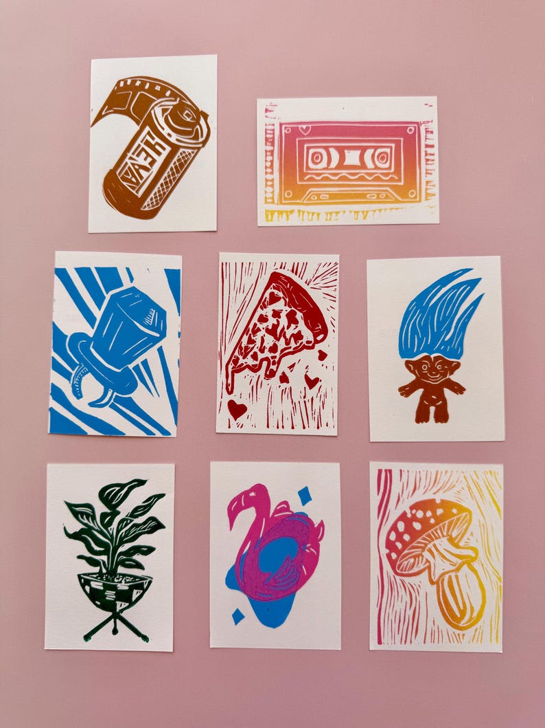 May include: A collection of linocut prints on white paper. The prints feature various designs, including a film canister, a cassette tape, a ring, a pizza slice, a troll doll, a potted plant, a flamingo float, and a mushroom.