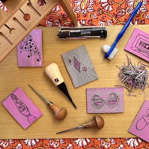 May include: A wooden cutting board displays various linocut tools and stamps. The stamps feature designs like a pizza slice, cassette tape, and heart-shaped sunglasses. A blue toothbrush and a graphite pencil are also present, along with a wooden tool holder.