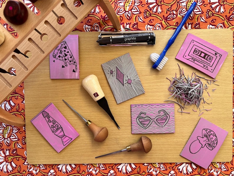 May include: A wooden cutting board displays various linocut tools and stamps. The stamps feature designs like a pizza slice, cassette tape, and heart-shaped sunglasses. A blue toothbrush and a graphite pencil are also present, along with a wooden tool holder.