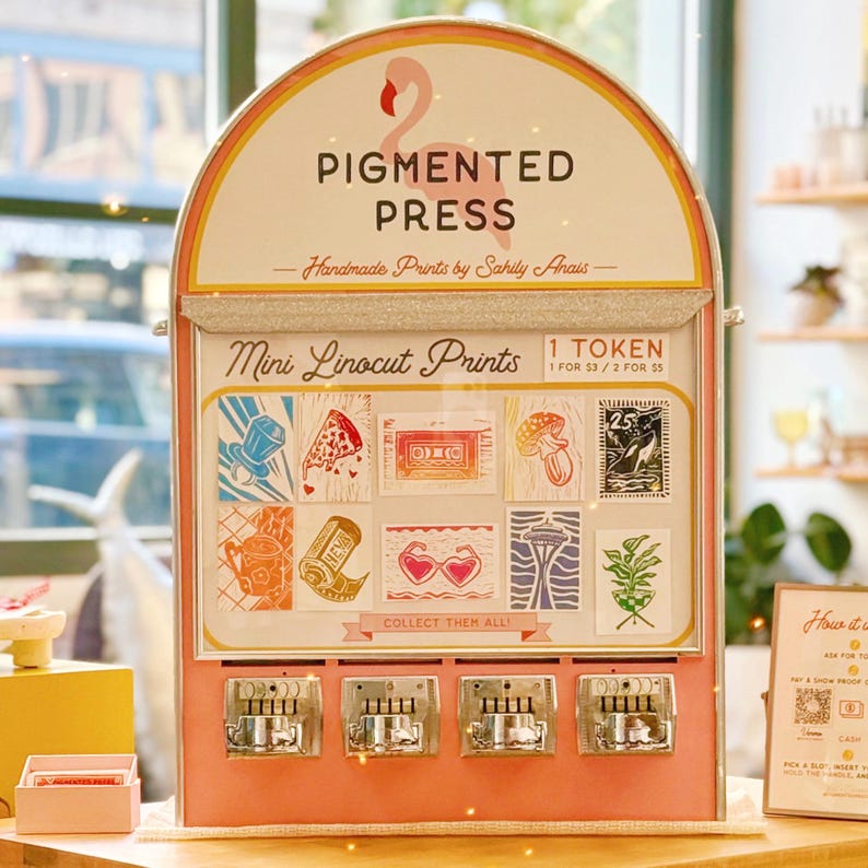 May include: A pink and gold vending machine with the text "PIGMENTED PRESS" and "Mini Linocut Prints." The machine dispenses small art prints with various designs, including a pizza slice and a cassette tape. The machine has a flamingo logo.