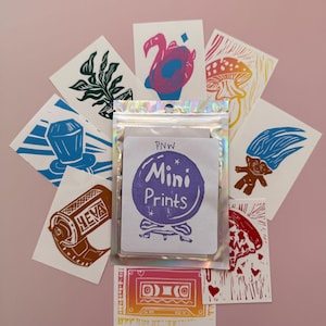 May include: A collection of mini prints featuring various designs, including a flamingo, mushroom, and cassette tape. The prints are arranged around a package labeled "PNW Mini Prints" on a pink background. The prints are in various colors.