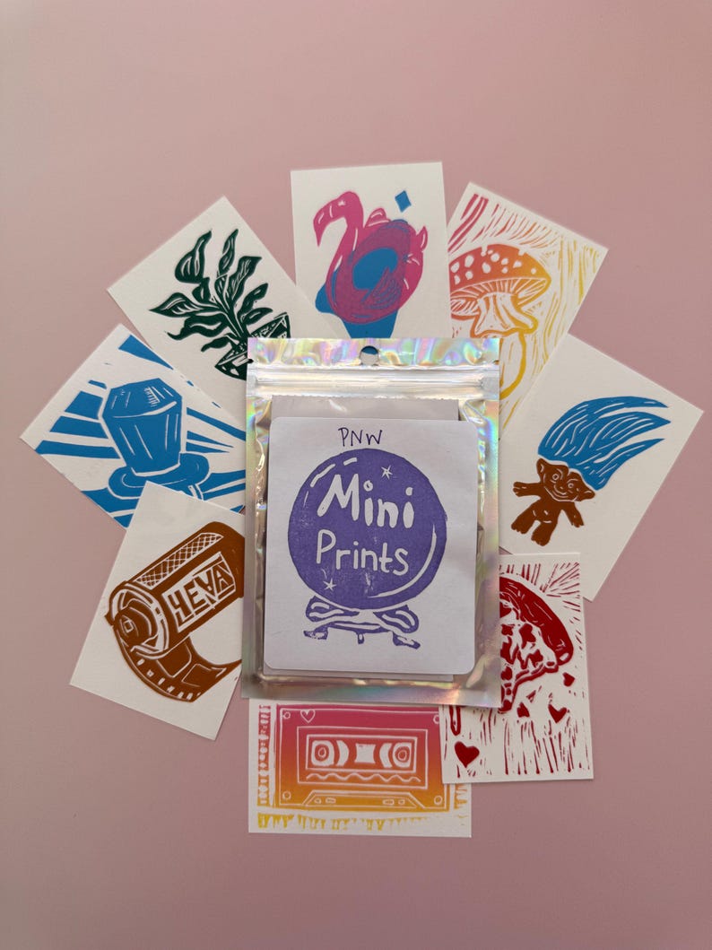 May include: A collection of mini prints featuring various designs, including a flamingo, mushroom, and cassette tape. The prints are arranged around a package labeled "PNW Mini Prints" on a pink background. The prints are in various colors.