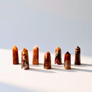 May include: Seven polished stone towers arranged in a row, each featuring unique banded patterns in shades of orange, brown, red, and cream. These vertical, faceted mineral points display natural variations in color, translucency, and dark inclusions.