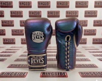 May include: Pair of iridescent boxing gloves with the "CLETD REYES PROFESSIONAL" logo. The gloves have a metallic sheen, with gold-colored laces and wristbands. The background features a repeating pattern of "RINGSIDE PRESTIGE" logos.