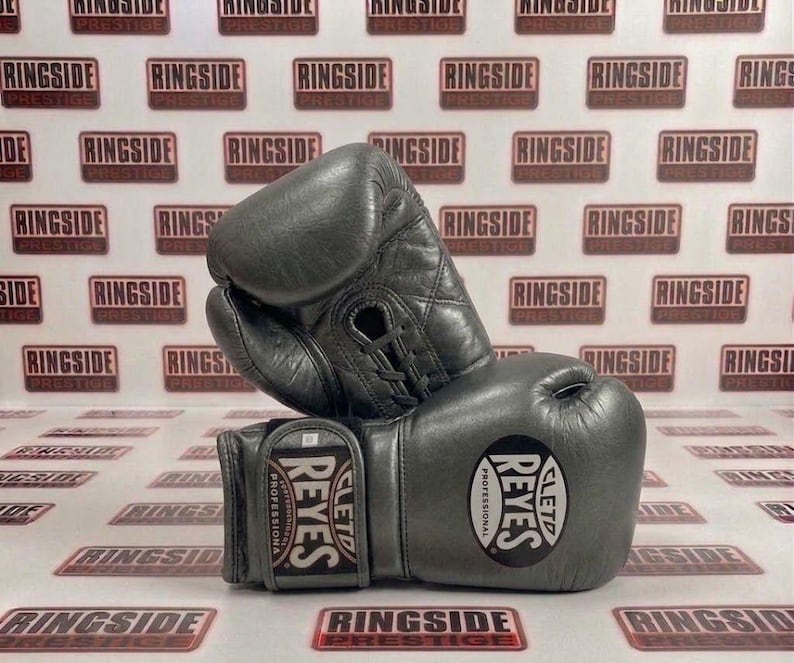 May include: A pair of gray leather boxing gloves, featuring the "REYES" logo and the words "PROFESSIONAL" and "CLETO". The gloves are laced up and appear to be new. The background has a repeating pattern of the word "RINGSIDE".