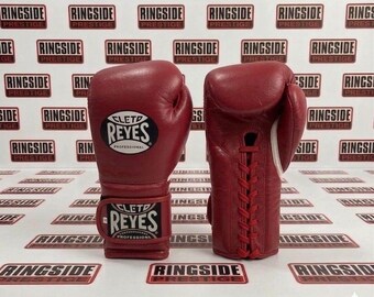 May include: Two red leather boxing gloves. One glove is upright, showing the lace-up closure on the back. The other glove is facing forward, displaying the brand name "CLETD REYES" in white lettering on a black background.
