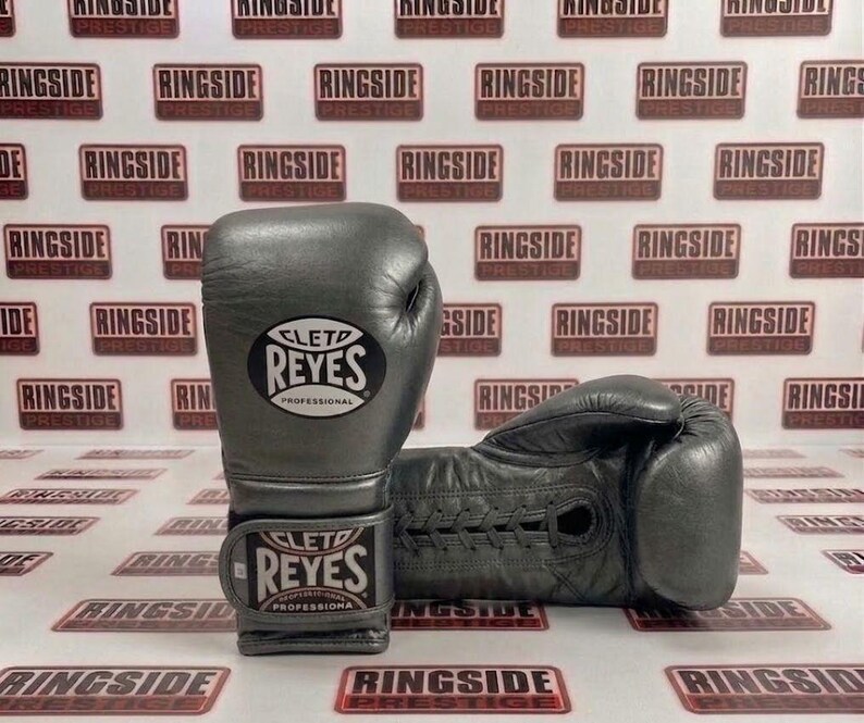 May include: A pair of gray leather boxing gloves with the brand name "CLETD REYES PROFESSIONAL" visible. The gloves are displayed on a background with the text "RINGSIDE PRESTIGE" repeated.