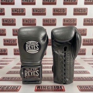May include: A pair of gray leather boxing gloves. The gloves are laced up and feature the "CLETO REYES PROFESSIONAL" logo. The background has a repeating pattern of "RINGSIDE PRESTIGE" logos.