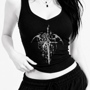 Puede incluir: Una camiseta de tirantes negra acanalada con cuello en V que presenta un estampado gráfico blanco de una espada rodeada de estrellas y formas aladas. La prenda se combina con pantalones de chándal negros con cordón y un collar de cadena plateada.