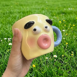 Puede incluir: Una taza de cerámica amarilla caprichosa con un diseño de cara de sorpresa. La taza presenta detalles negros para los ojos y las orejas, una boca rosa y un asa azul claro. La taza se sostiene en una mano sobre un fondo de hierba verde.