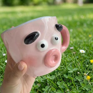 Puede incluir: Una taza de cerámica rosa con diseño de cara de cerdo. La taza presenta detalles negros para los ojos y las orejas, un hocico rosa y un asa a juego. La taza se sostiene sobre un fondo de hierba verde.