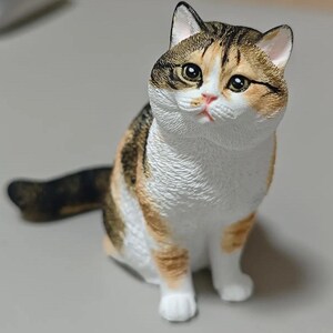 Puede incluir: Figura detallada de un gato sentado con un pelaje tricolor marrón, naranja y blanco. El gato tiene ojos grandes y expresivos y una nariz rosada. La cola está enrollada hacia un lado. Es un objeto decorativo.