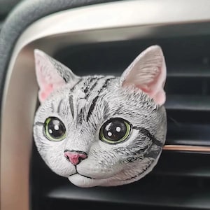 Könnte beinhalten: Ein grauer und weißer Katzenkopf-Auto-Luftauslassclip. Die Katze hat große grüne Augen, eine rosa Nase und rosa Innenohren. Das Fell der Katze hat schwarze Streifen. Der Clip ist an einem Lüftungsschlitz befestigt.