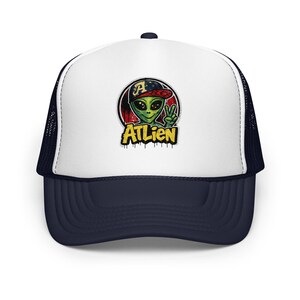 May include: A white and navy blue trucker hat featuring a graphic of a green alien wearing a baseball cap with the letter A, making a peace sign. Below the alien, the text ATLIEN is printed in yellow with a black outline and drip effect.