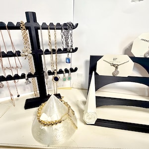 Pode incluir: Exibição de colares e pulseiras em expositores pretos. Vários colares com diferentes pingentes e estilos de corrente estão pendurados em um expositor em camadas. Uma pulseira dourada é exibida em um suporte branco em forma de cone. Outros colares são exibidos em cartões brancos.