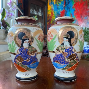 May include: A pair of ceramic vases featuring a cream-colored base with hand-painted figures in blue, red, and green robes. Each vase displays a gold-rimmed opening, a black base, and intricate floral accents surrounding the central character design.
