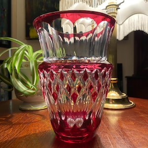Pode incluir: Um vaso de cristal transparente com borda e base vermelho rubi. O vaso apresenta um padrão lapidado em diamante na metade inferior e um design liso e curvo na metade superior. O vaso é provavelmente usado para fins decorativos.