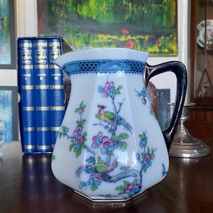 Op de afbeelding: Witte porseleinen kan met een zwarte handgreep en een bloemen- en vogelontwerp. De kan heeft een blauwe rand en een gouden voet. Het ontwerp bevat kleurrijke vogels en roze en blauwe bloemen op takken.