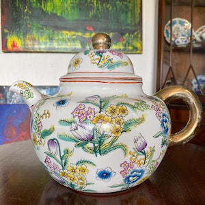 May include: A white ceramic teapot featuring a colorful floral pattern with yellow, purple, and blue flowers and green foliage. The teapot has a gold-colored handle, a matching lid with a gold-toned knob, and a short spout with floral details.