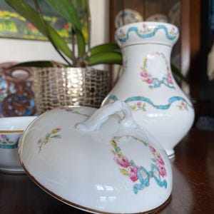 May include: A white porcelain lidded vessel and a matching vase, both featuring a delicate pattern of pink roses, green leaves, and light blue ribbons. The lid has a curved handle, and both pieces are finished with a thin gold rim detail.