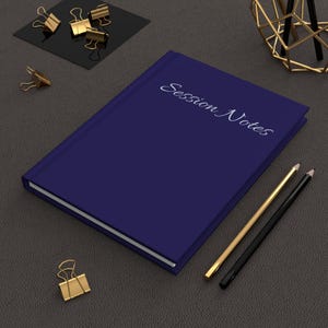 May include: A navy blue hardcover journal with the words "Session Notes" in cursive script. Gold and black pencils and gold binder clips are arranged around the journal on a dark gray surface.