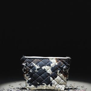 May include: A quilted pouch featuring a black and white cow print pattern. The rectangular bag has a zippered top closure and a structured base, showcasing a distinct diamond-stitched texture across the exterior surface of the fabric.
