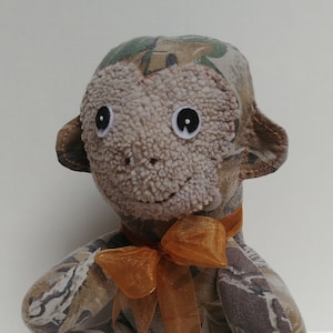 May include: A brown and green camouflage monkey plush toy with a brown fuzzy face and large black eyes. The monkey is wearing a brown and orange ribbon around its neck.