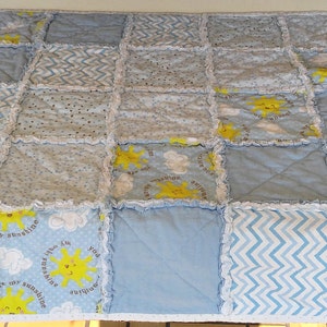 May include: A baby quilt made of blue and white fabric with a patchwork design. The quilt features a variety of patterns, including stars, clouds, and zigzags. The quilt is edged with white lace.