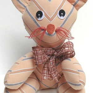 May include: A handmade plush cat toy with a striped fabric pattern in shades of peach and blue. The cat has a red nose, black eyes, and a brown and white plaid bow.
