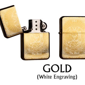 May include: Gold-colored lighter with white engraving, featuring an open and closed view. The design includes a sunburst pattern and an illustration of a devil, with the text "The Devil" at the bottom. The word "GOLD" is displayed below.