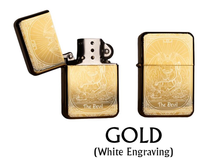 May include: Gold-colored lighter with white engraving, featuring an open and closed view. The design includes a sunburst pattern and an illustration of a devil, with the text "The Devil" at the bottom. The word "GOLD" is displayed below.