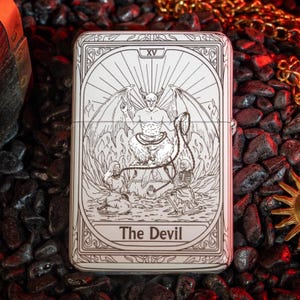 May include: A silver-colored lighter featuring a detailed engraving of The Devil tarot card. The design includes a horned figure with wings, surrounded by figures and the text "The Devil". The lighter has a rectangular shape.