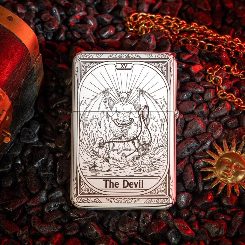 May include: A silver-colored lighter featuring a detailed engraving of The Devil tarot card. The design includes a horned figure with wings, seated above two figures and a skeleton. The text "The Devil" is at the bottom.