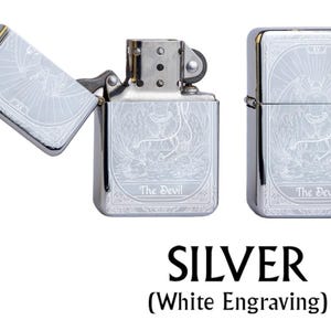 May include: Silver lighter with white engraving featuring a detailed illustration of The Devil tarot card. The lighter is shown open and closed, with the text "The Devil" at the bottom. The word "SILVER" is displayed below the lighters.