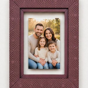May include: A claret-coloured picture frame with a geometric pattern surrounds a family portrait. The frame has a textured surface and a rectangular shape. The photograph features a smiling family of four.