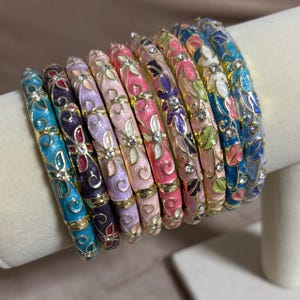May include: A collection of nine metal bangle bracelets featuring colorful enamel floral patterns and embedded clear rhinestones. Each bracelet displays a unique color palette including shades of blue, purple, pink, and white with gold-tone metal accents.