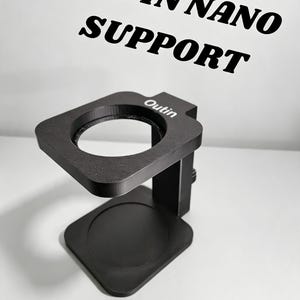 Puede incluir: Un soporte negro Outin Nano con base cuadrada, columna vertical y plataforma superior con abertura circular. La palabra Outin está impresa en blanco en la plataforma y el texto OUTIN NANO SUPPORT aparece en la parte superior.