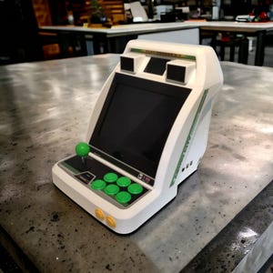 May include: A white and green retro arcade console with a black screen, green joystick, and green buttons. The console has a sleek design with a curved top and side accents. The console is sitting on a gray surface.