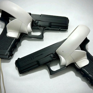 May include: Two black pistol-shaped controllers featuring white VR controller attachments mounted on the grips. The black frames have a textured handle and a trigger guard, with the white plastic VR components secured over the top and side.