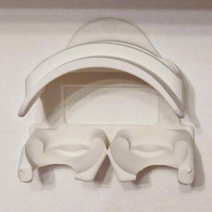 May include: A white wall-mounted plastic organizer featuring a curved top shelf and two lower compartments designed to hold items. The structure has a smooth, matte finish with a central rectangular recessed area and a mounting hole at the top.