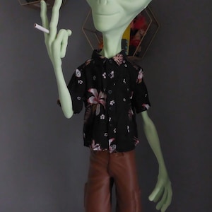 Peut inclure: Figurine d'extraterrestre vert portant une chemise noire à motifs floraux, un short marron et des tongs roses. L'extraterrestre tient une cigarette et fait le signe de la paix.