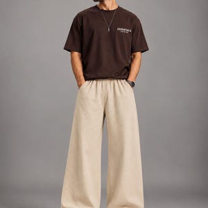 May include: A dark brown short-sleeved t-shirt with the text ESSENTIALS printed in white on the chest, paired with wide-leg beige trousers featuring an elastic waistband and side pockets, worn with white sneakers, a silver necklace, and sunglasses.