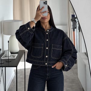 May include: A dark wash denim jacket featuring a mandarin collar, a front button closure with silver-toned buttons, two chest pockets, and voluminous sleeves with pleated detailing at the cuffs. The jacket is paired with matching dark denim jeans.
