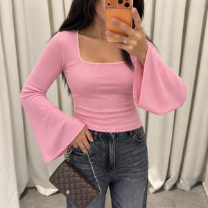 Peut inclure: Un haut rose à manches longues avec une encolure carrée ornée d'un liseré festonné blanc et des manches évasées, porté avec un jean en denim gris foncé et un petit sac à bandoulière rectangulaire en cuir marron matelassé.