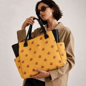 Peut inclure: Un sac cabas jaune moutarde orné d'un motif répétitif de petites grappes de fleurs rouges et vertes. Le sac comprend des anses en similicuir noir et des panneaux latéraux noirs, avec des poches latérales ouvertes sur les côtés gauche et droit.