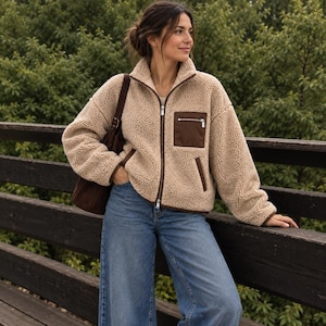 May include: A beige sherpa fleece jacket with a high collar, a full-length front zipper, and long sleeves. The jacket features a dark brown chest pocket with a zipper closure and dark brown trim along the zipper and pocket edges.