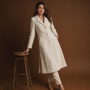 May include: A long, cream-coloured coat featuring a notched lapel, long sleeves, and a three-button front closure. The coat is paired with matching cream-coloured trousers and tan pointed-toe shoes, creating a coordinated ensemble with a structured fit.