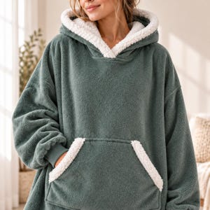 May include: An oversized hooded sweatshirt in a muted sage green color, crafted from a textured fleece material. The garment features a thick white sherpa lining inside the hood and matching white sherpa trim on the large front kangaroo pocket.